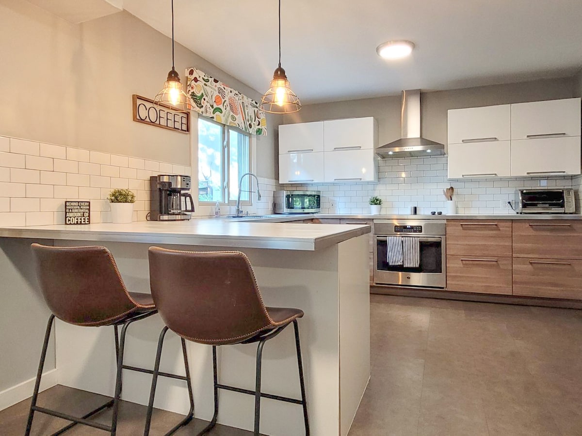 A modern kitchen features sleek cabinetry in light and wood tones, equipped with stainless steel appliances. A central island with two chairs provides a casual dining option, while natural light enters through a nearby window with floral-patterned trim.