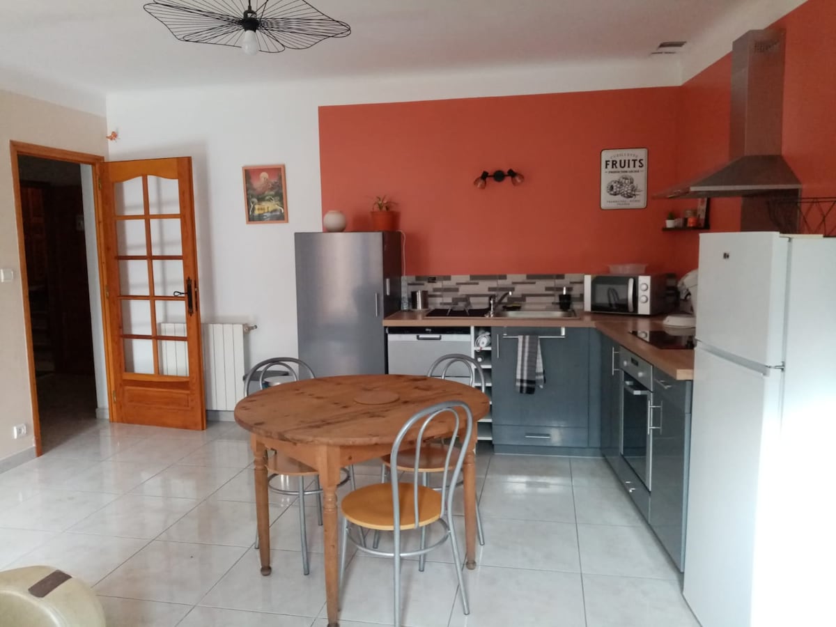 A well-equipped kitchen and dining area feature a round wooden table with three metal chairs. Grey cabinets and modern appliances, including a refrigerator and microwave, complement the coral wall. A window allows natural light to enhance the spacious setting.