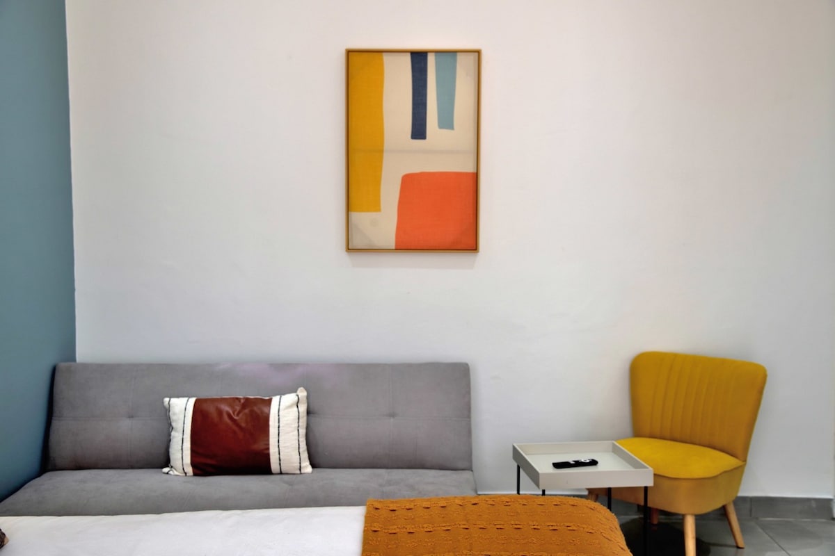 A lounge area features a light gray sleeper couch adorned with a decorative pillow. A small table and a mustard-yellow chair are positioned alongside the couch. A framed abstract artwork adds a touch of color to the white wall.