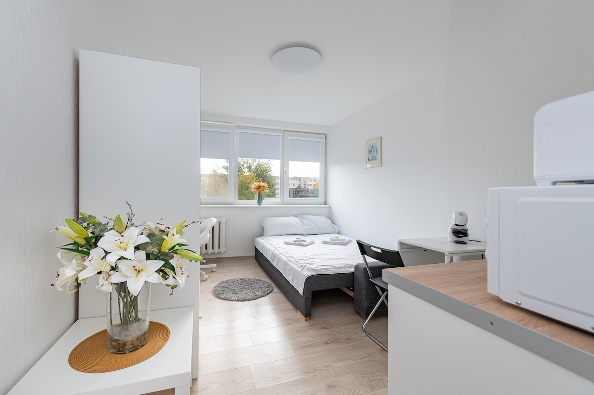 A minimalistic room features a spacious bed with fresh linens, positioned near large windows that allow ample natural light. A small table with a chair and a microwave are present, alongside a vase of flowers atop a white sideboard, adding a touch of color.