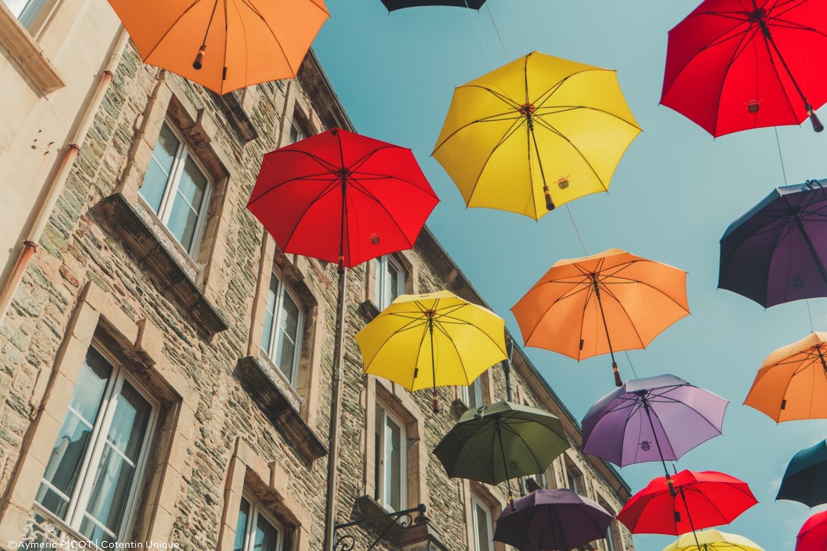 Colorful umbrellas in shades of red, yellow, purple, and orange are suspended above a charming street. The background features a stone building with classic windows, providing a vibrant contrast to the umbrellas against the clear blue sky.