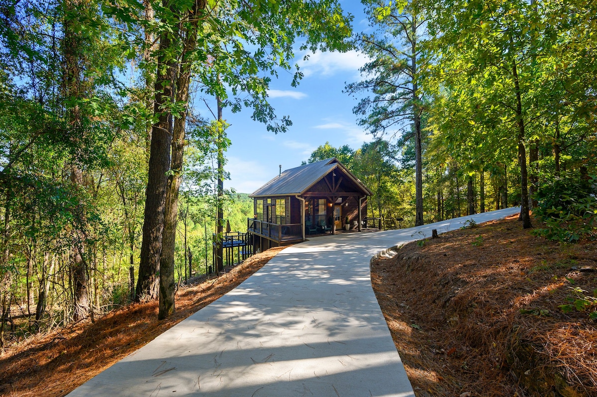 The cabin is situated among lush greenery, with a paved driveway leading to the entrance. Tall trees frame the scene, creating a peaceful setting in nature. The structure features a welcoming front porch, inviting guests to relax and enjoy the surrounding forest.