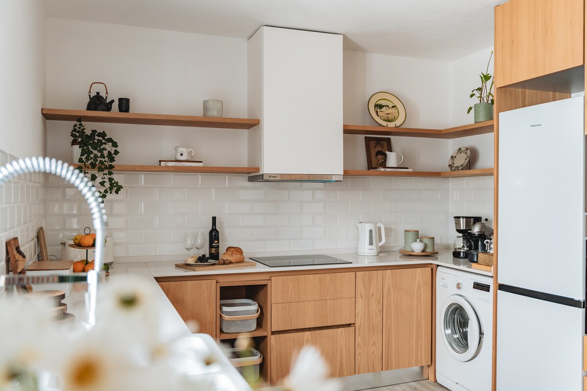 A modern kitchen features wooden cabinets and open shelving. A white countertop is complemented by a glass-ceramic cooktop and an array of kitchen appliances. Natural light illuminates the space, highlighting the thoughtful organization with pots, utensils, and decorative items on display.