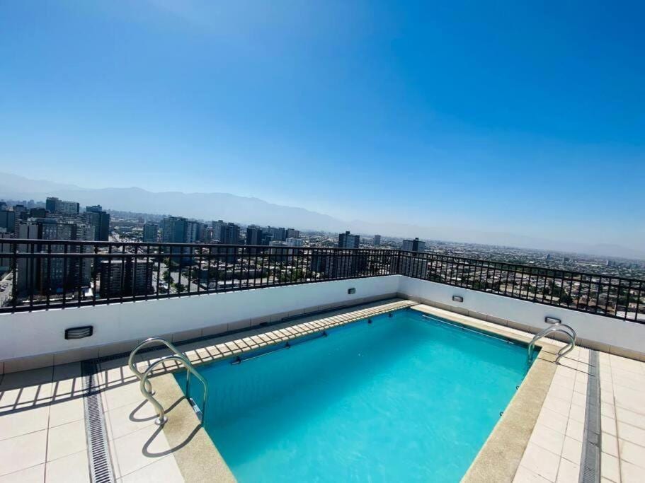 A rooftop pool is presented, featuring clear blue water and a surrounding terrace. A railing provides safety while offering expansive views of the cityscape and mountains in the background. The sky is bright and clear, highlighting the inviting space.