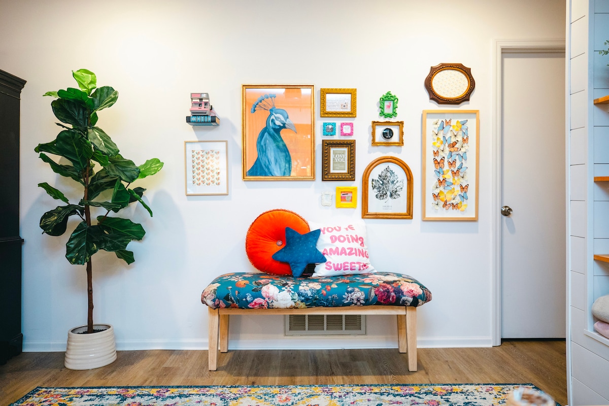 A cozy entryway features a colorful bench adorned with floral upholstery and vibrant cushions. A collection of framed artworks and decorative items hangs on the wall behind the bench, complemented by a tall potted plant. Natural light enhances the welcoming atmosphere.