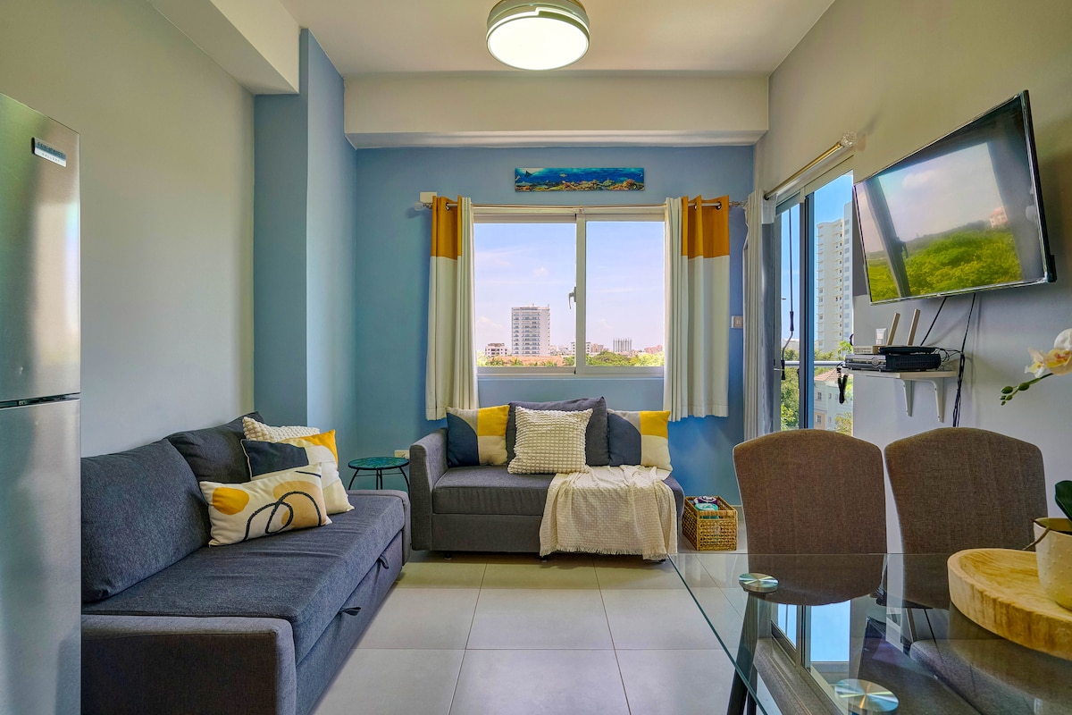 A modern living area features a comfortable gray sofa and a smaller loveseat, accented with cushions. A glass dining table is visible alongside, with four chairs. Natural light enters through a large window, framed by yellow and white curtains, offering views of the surrounding area.