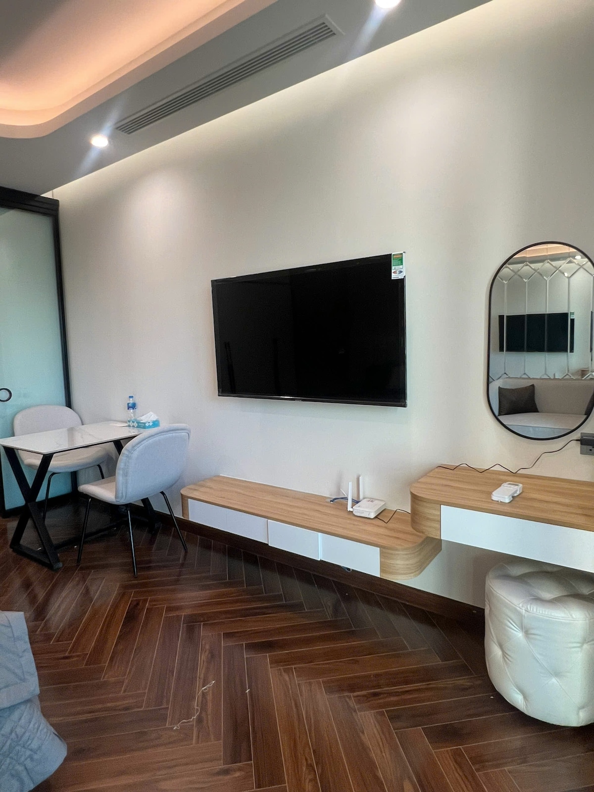 A well-furnished studio apartment features a large wall-mounted TV opposite a small dining table with two chairs. A sleek console table is positioned beneath an elegant mirror, and the warm wooden flooring adds to the cozy ambiance.