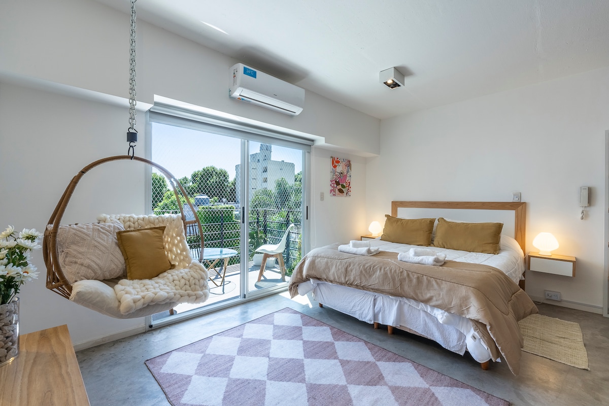 A bright sleeping area features a spacious bed with neutral-toned linens and decorative pillows. A hanging chair is positioned nearby. Large glass doors provide access to a balcony, allowing natural light to fill the room. A geometric rug adds a subtle touch of texture.