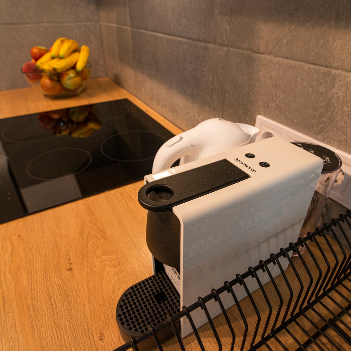A modern coffee machine is positioned on a wooden countertop beside a sleek stovetop. A bowl of fresh fruit, including bananas and apples, is visible in the background, highlighting a functional kitchen setup.