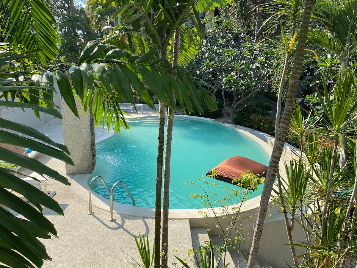 The serene swimming pool is nestled among tropical greenery, featuring crystal clear water. A round sunbed can be seen partially submerged, offering a relaxing spot beside the pool. Surrounding plants contribute to a calm and natural ambiance.