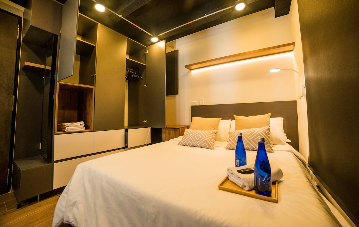 The bedroom features a spacious bed with two decorative pillows and a light blanket. A wooden tray with two blue bottles and towels rests on the bed. Modern storage units are positioned along the walls, providing ample space for belongings.