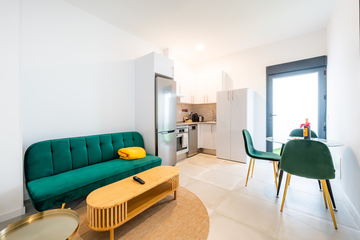 A modern living space features a green sofa with a yellow throw, complemented by a wooden coffee table. A dining table with green chairs sits nearby. Stainless steel appliances and white cabinetry create a clean, functional kitchen area. Natural light enters through a door leading to the outside.