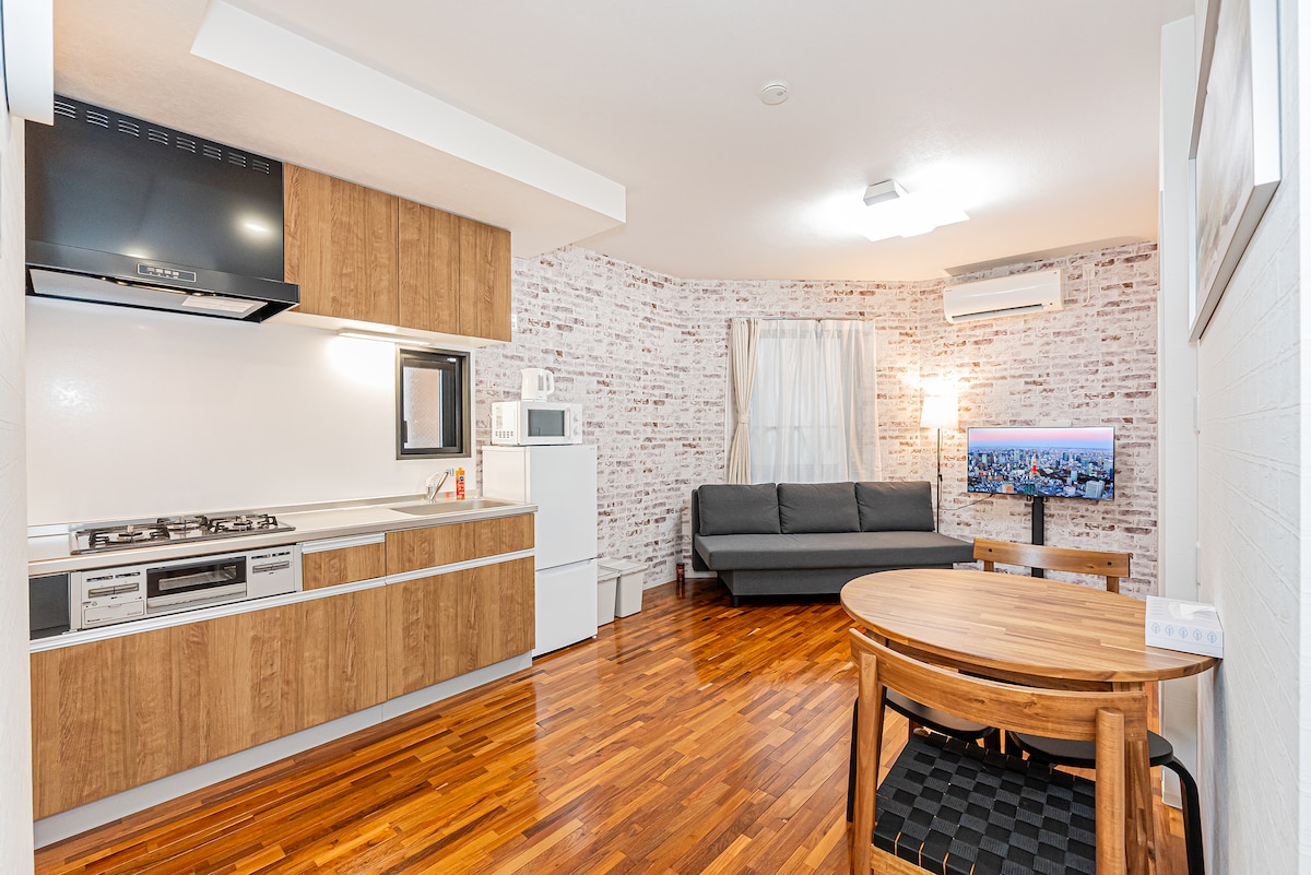 The open-plan living area combines a kitchen and seating space, featuring modern wooden cabinetry and a stylish countertop. A gray sofa offers comfortable seating, and a table with wooden chairs creates a dining area. A large window allows natural light to fill the space.
