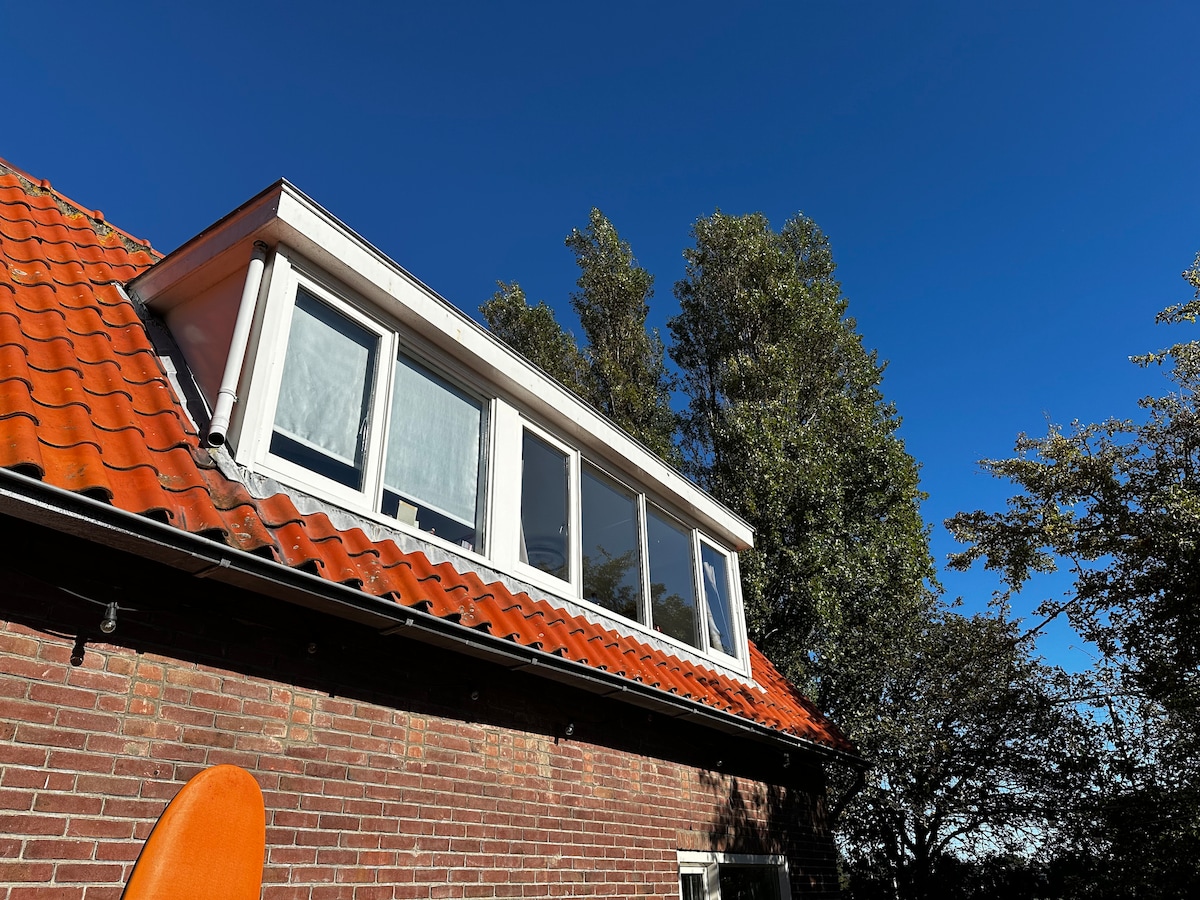 The image captures the upper part of a cozy holiday home. The red tile roof complements the brick exterior. Large windows provide a view into the upper space, while surrounding trees enhance the setting against a clear blue sky.