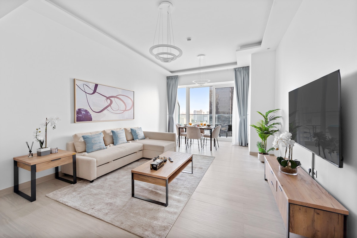 A spacious living area features a comfortable sofa adorned with pillows, a stylish coffee table, and a smart TV mounted on the wall. Large windows allow natural light to fill the space, with a dining table visible in the background and potted plants adding greenery.