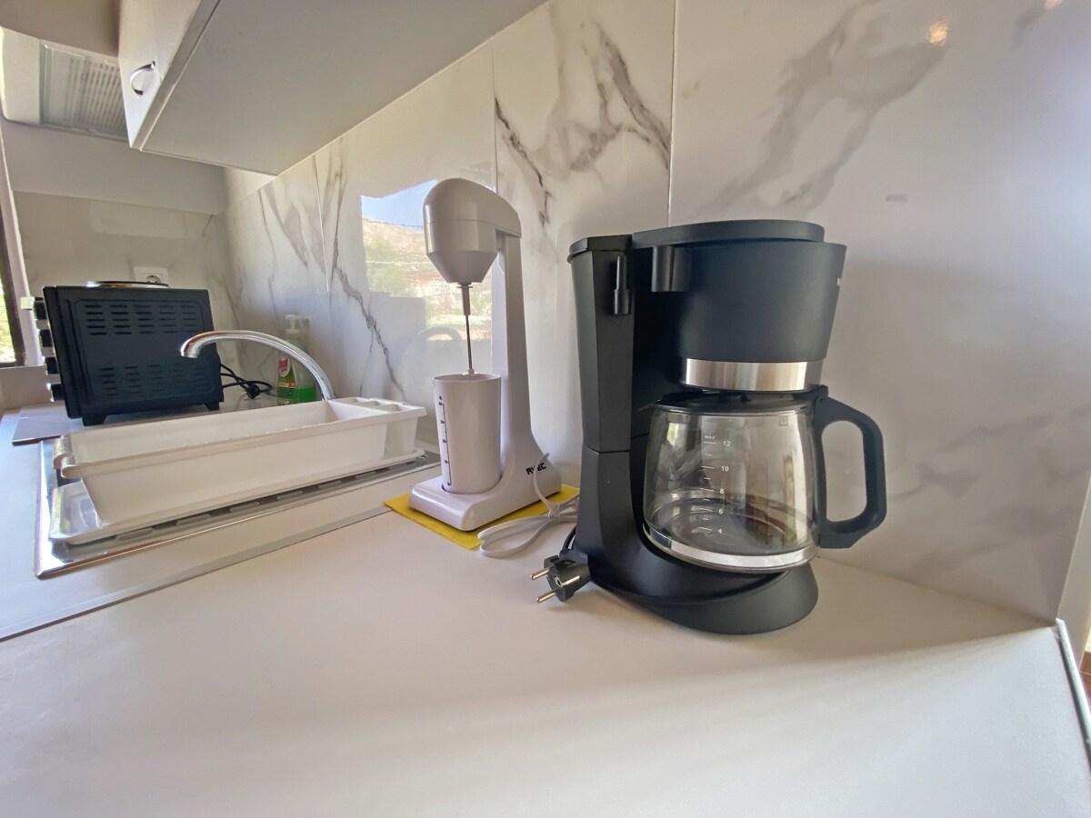 A coffee maker with a glass carafe is positioned on a counter near a blender and a drying rack. The countertop has a smooth, light-colored surface, complemented by a marble-patterned backsplash that adds a modern touch to the kitchen space.