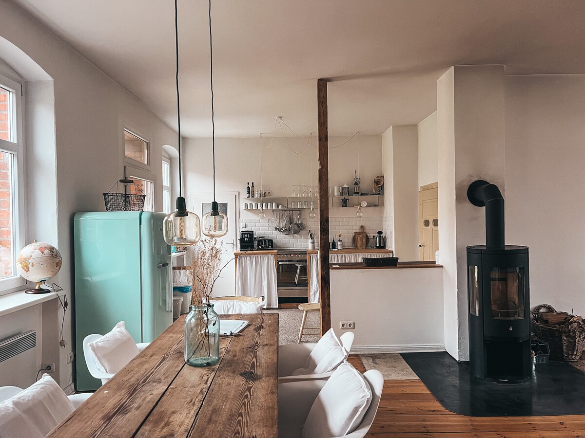 A modern kitchen and dining area feature a wooden table surrounded by four chairs. A retro fridge in mint green contrasts with white walls. Shelves hold kitchen essentials. A corner wood stove adds warmth to the space, which is softly illuminated by hanging pendant lights.