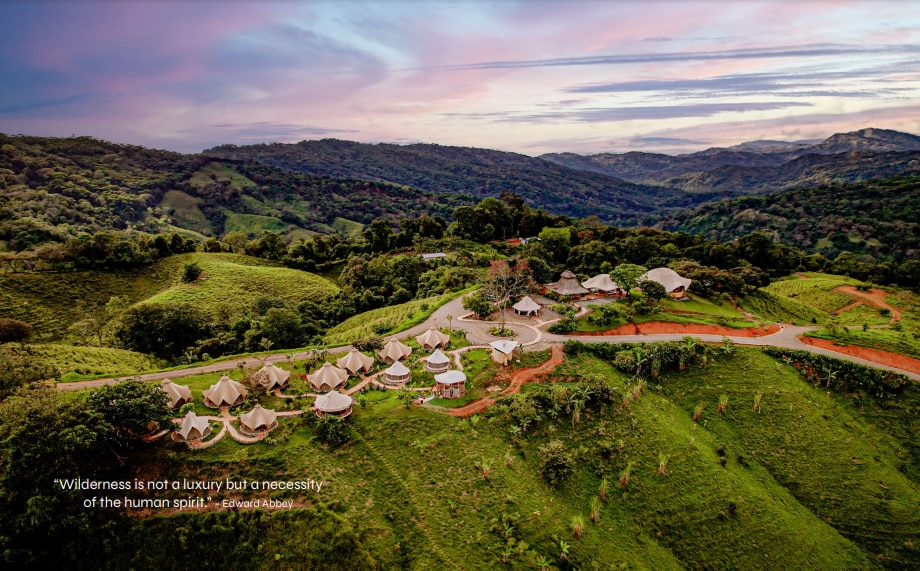 An aerial view captures the layout of glamping tents nestled among rolling hills in a lush green landscape. The setting is surrounded by dense trees and showcases a winding road that leads through the area, illustrating a harmonious blend of nature and accommodation.