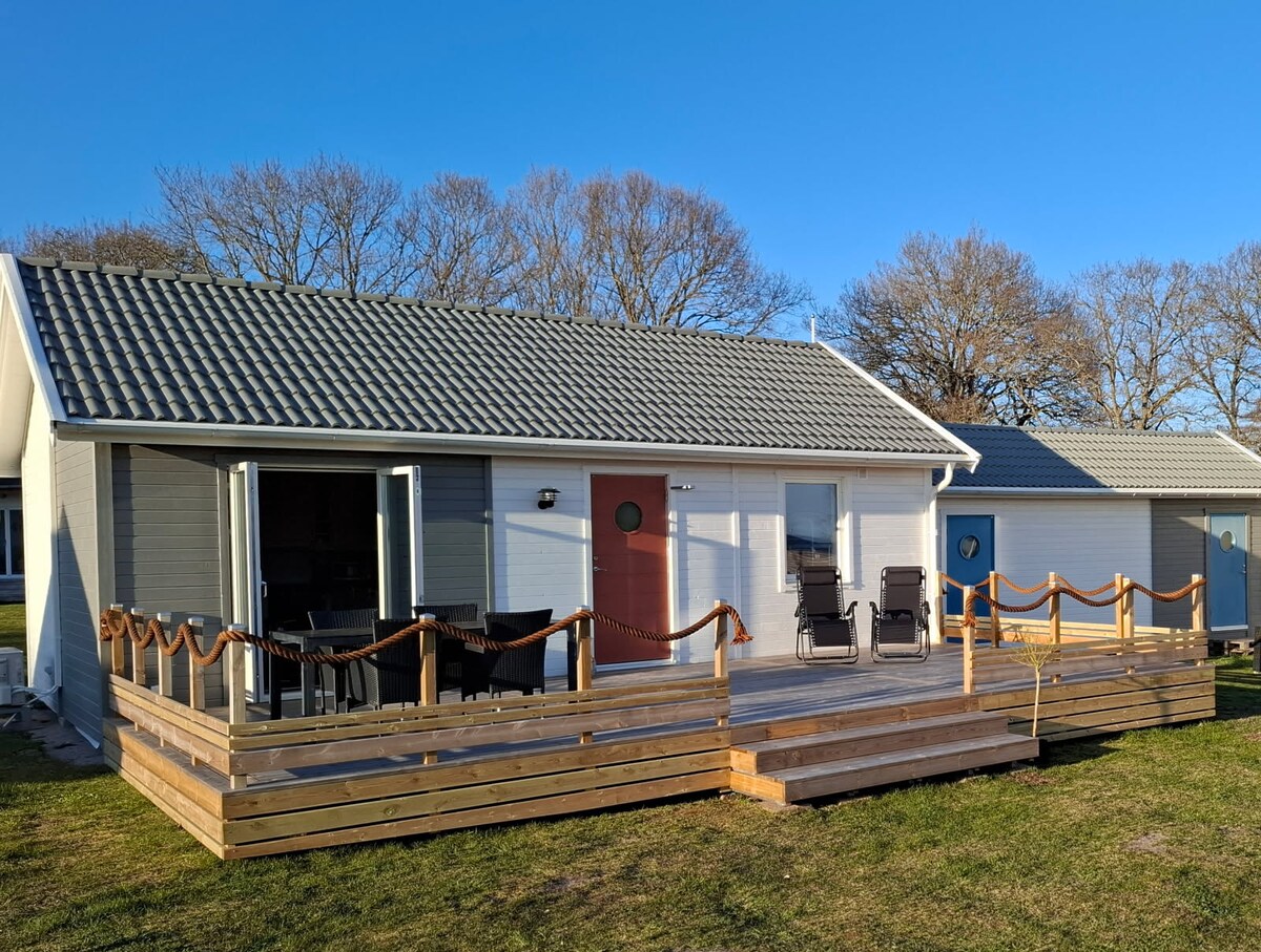 A modern exterior of a family-friendly building is displayed, featuring a gray roof and a welcoming red door. Surrounding greenery complements the wooden deck, which has a railing and is furnished with several chairs positioned for outdoor relaxation.