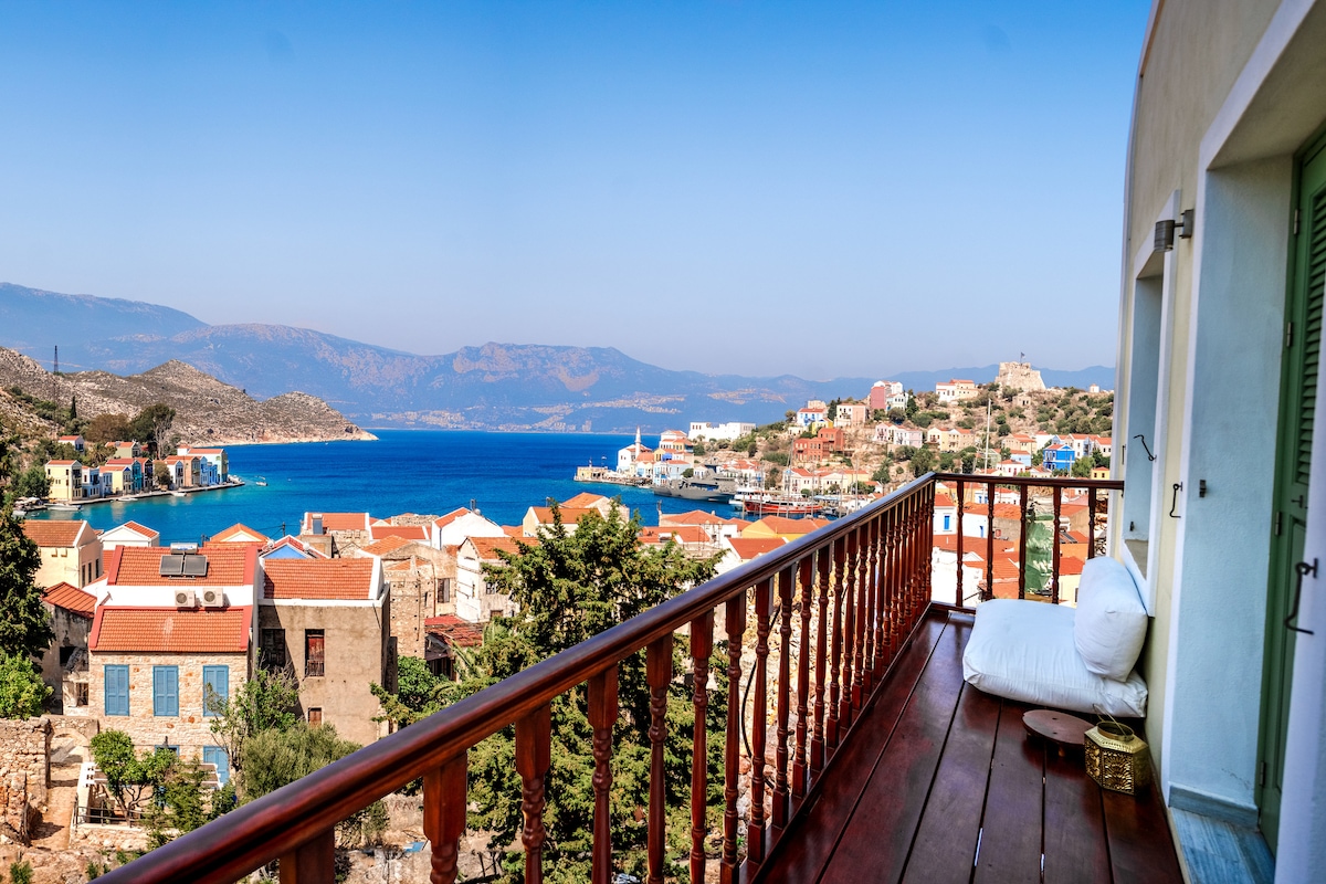 A balcony view captures a vibrant coastal town with colorful buildings nestled against the water. The vast blue sea meets the sky in the distance, while mountains frame the landscape. A large cushion is placed invitingly on the wooden railing, enhancing the serene setting.