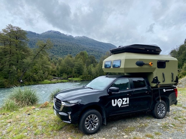 A Mazda BT 50 4x4 camper is parked beside a tranquil river, surrounded by lush greenery and mountains. The roof tent is visible, offering additional sleeping space. The environment reflects a connection with nature, ideal for outdoor adventures.