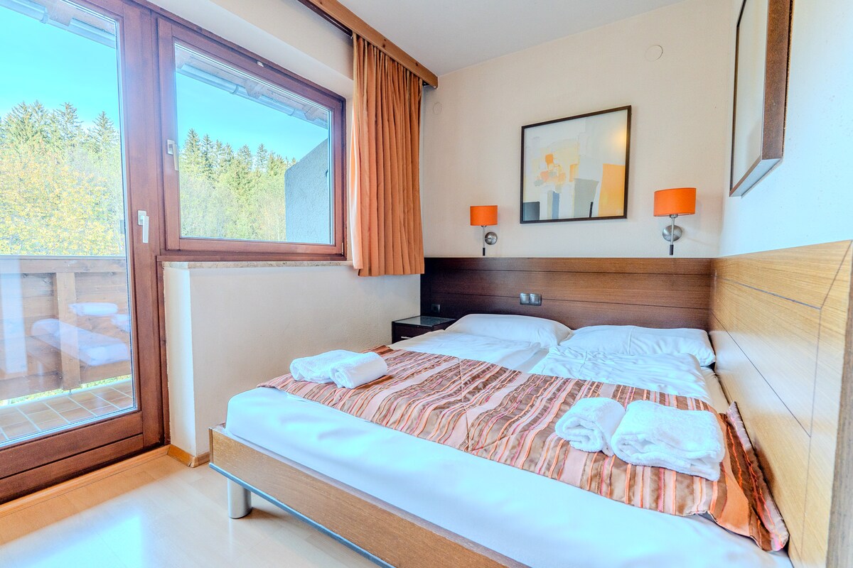 A cozy bedroom is arranged with a spacious bed featuring a striped blanket and neatly placed towels. Warm orange lamps are mounted on the wooden headboard, complementing the warm tones of the room. A balcony door is visible, leading to a view of greenery.