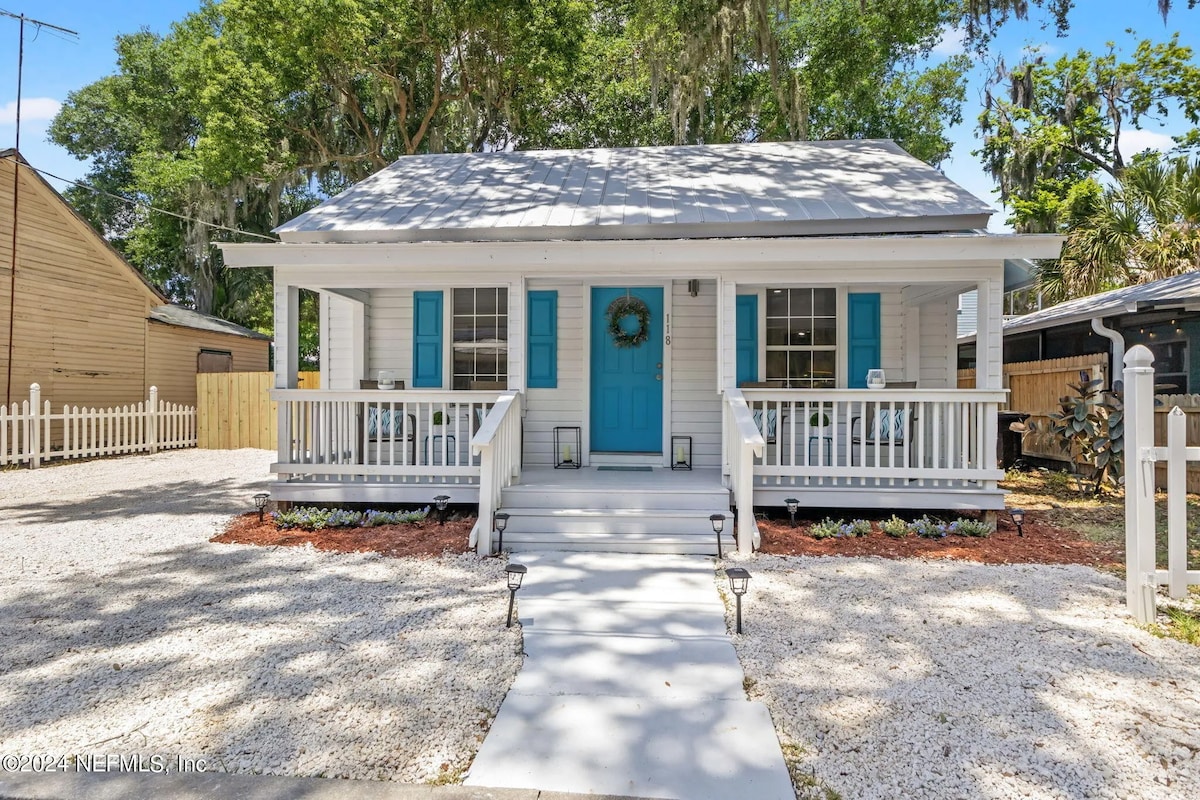 A charming bungalow exterior is showcased, featuring a light-colored facade and blue shutters. A welcoming front porch with white railings is visible, accompanied by a white pathway leading to a blue front door. Surrounding greenery adds to the inviting atmosphere.