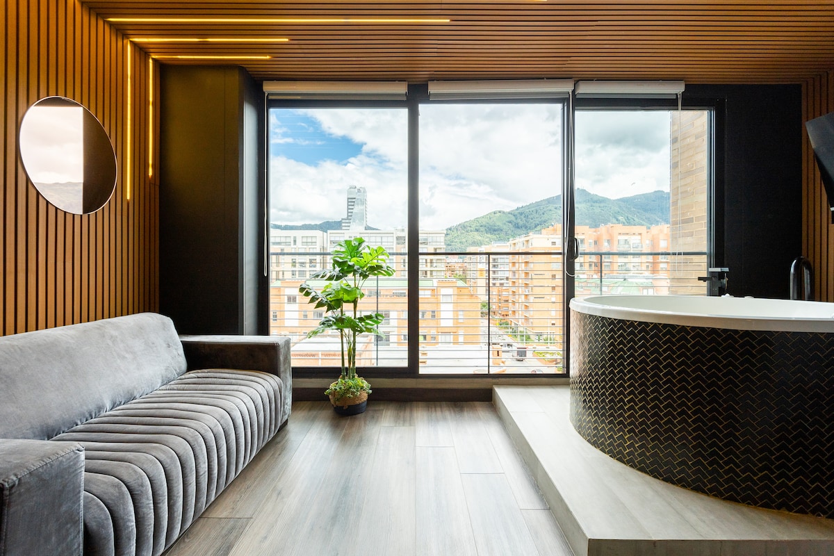 A modern living space features a plush gray sofa positioned beside a circular jacuzzi. Large windows frame a cityscape view, with distant mountains visible in the background. Natural light fills the room, highlighting wood accents and sleek finishes.