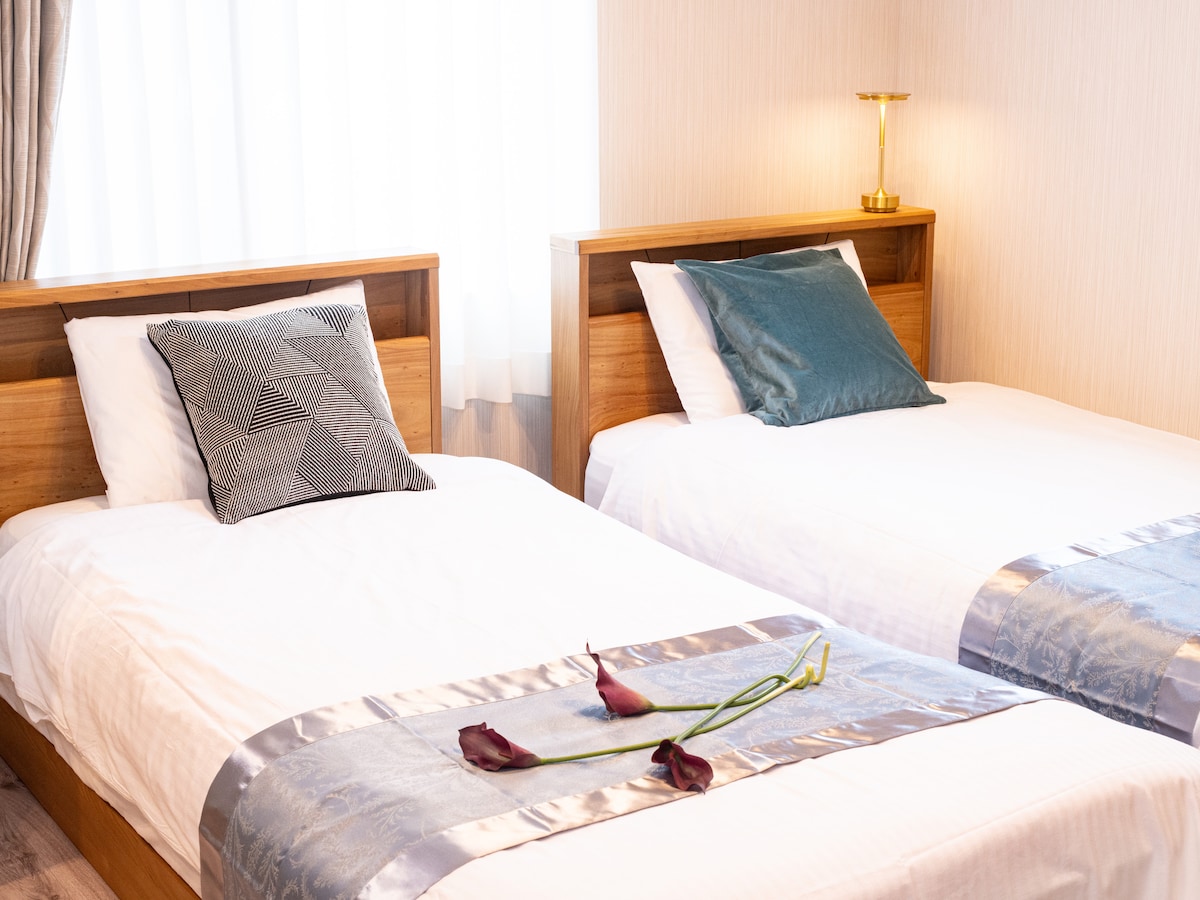 The sleeping area features two spacious beds, each adorned with decorative pillows. A soft, light-colored blanket complements the bedding. Fresh flowers are elegantly placed on the bedspread, while natural light filters in through sheer curtains, creating a serene atmosphere.