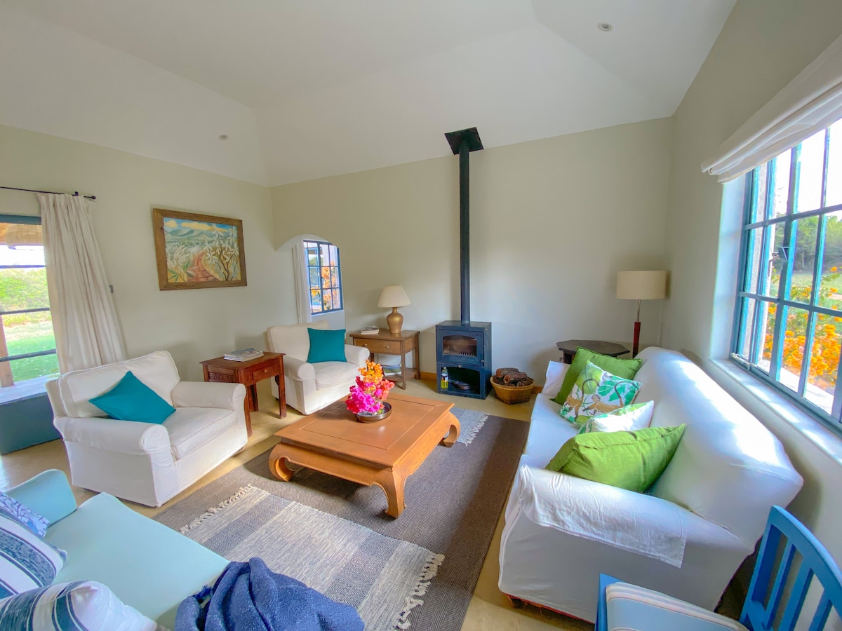 A welcoming living room is highlighted by comfortable white sofas adorned with green and blue cushions. A wooden coffee table sits in the center, decorated with vibrant flowers. A fireplace stands against a pale wall, and large windows allow natural light to fill the space.