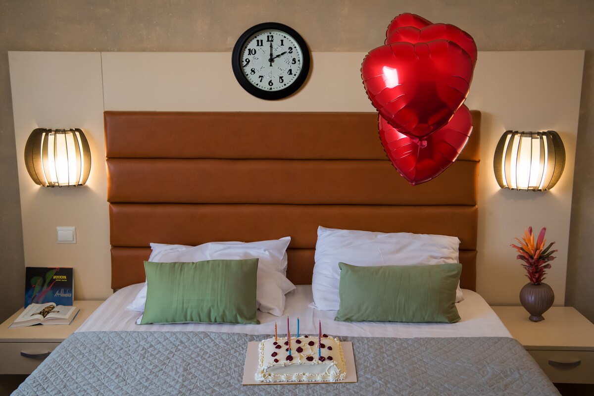A cozy double room is furnished with a comfortable bed, adorned with two green pillows. Above the bed, a clock is mounted on the wall, flanked by two stylish light fixtures. A cake sits on the bed, accompanied by red heart-shaped balloons, adding a celebratory touch.