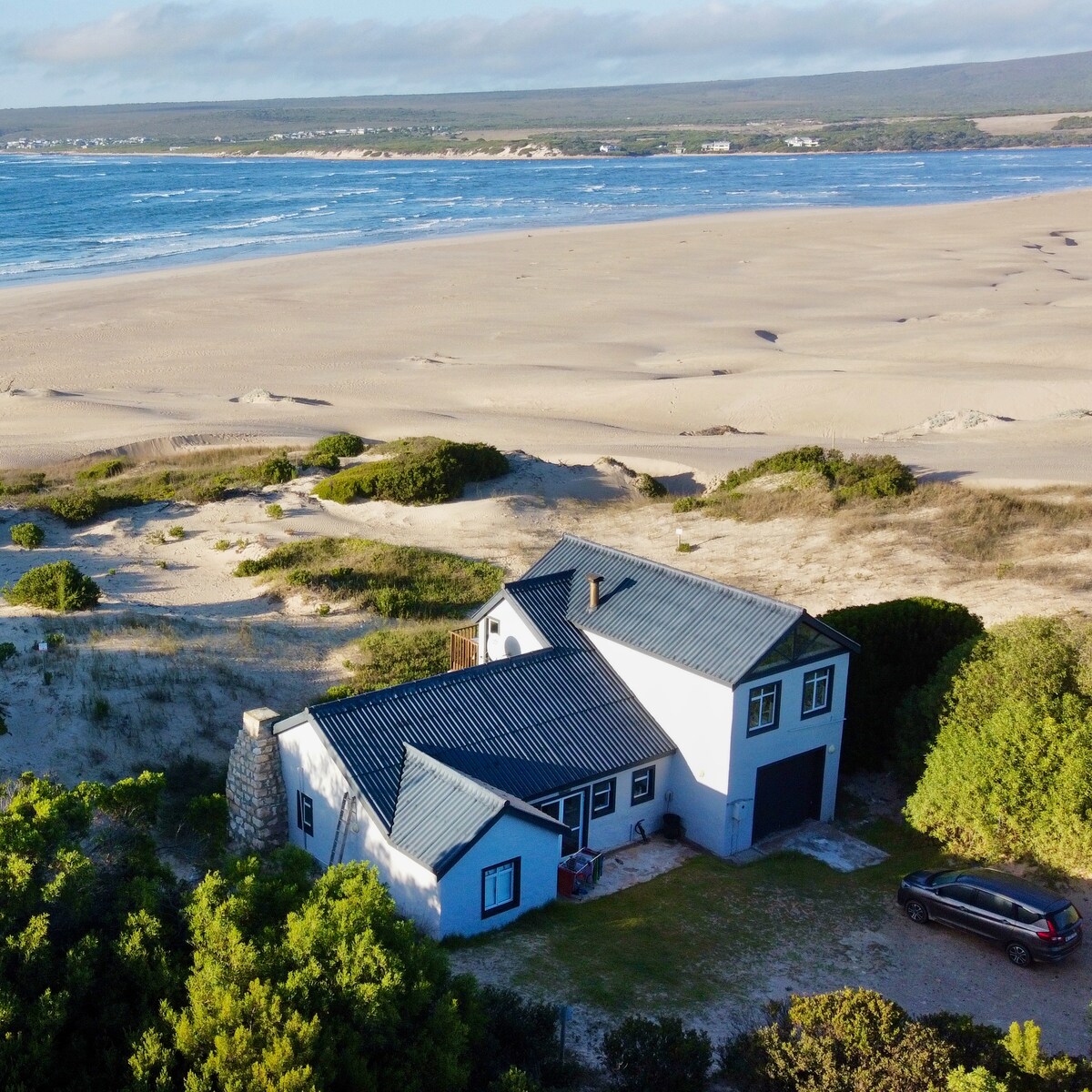The coastal beach house is surrounded by sand dunes, with expansive ocean views visible from the property. A vehicle is parked nearby, while landscaped greenery adds a natural touch to the landscape, creating a serene and secluded environment.