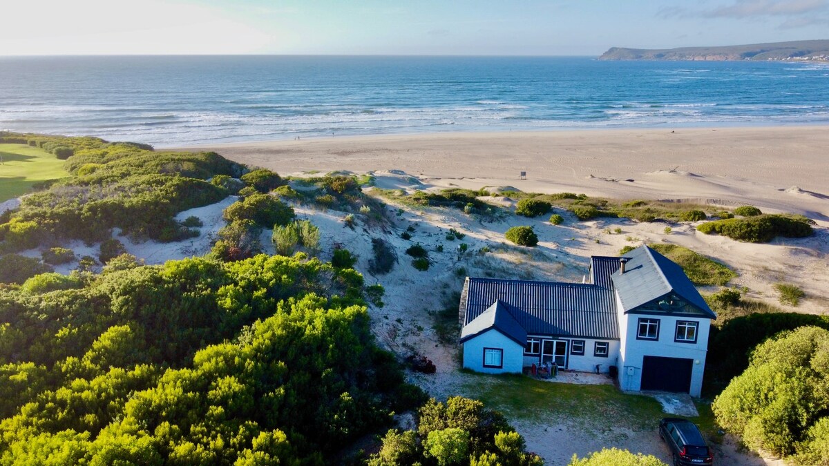 An aerial view showcases a beach house nestled amongst green dunes, with expansive sandy shores stretching toward the ocean. The house, featuring a dark roof and white walls, is surrounded by lush vegetation, emphasizing its serene coastal setting.
