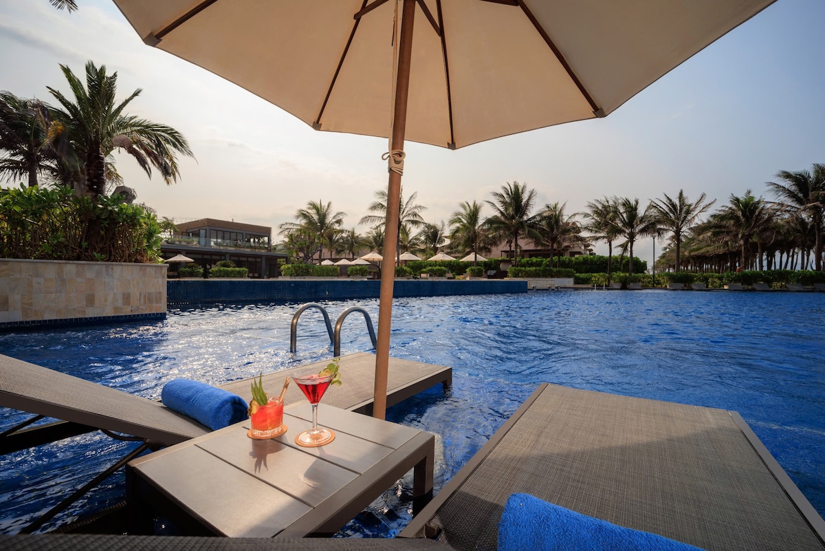 A luxurious pool area is featured, with clear blue water and lush palm trees surrounding the space. Two beverages sit on a table beside a lounge chair, offering a relaxing atmosphere under a large umbrella.