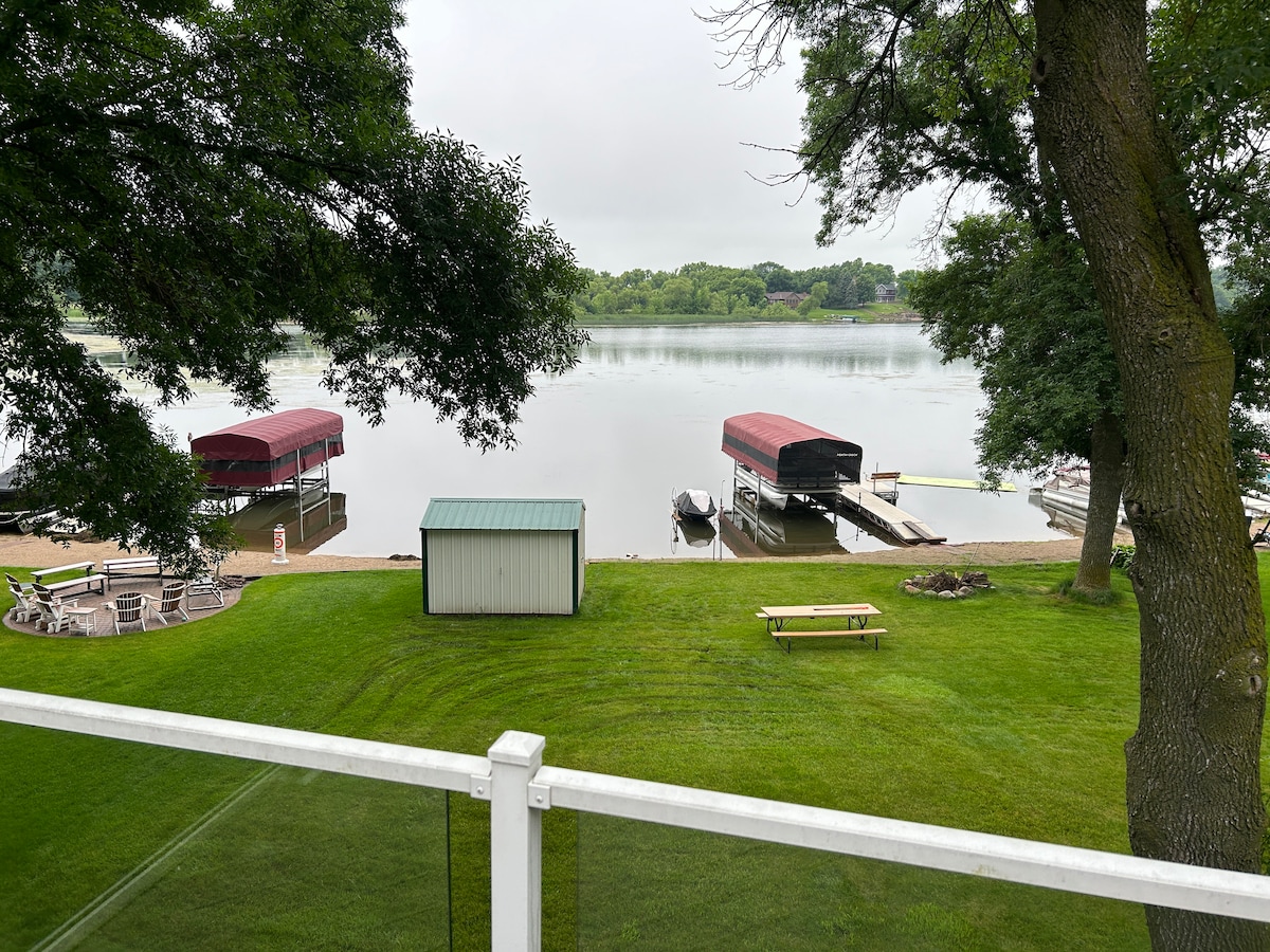 A view overlooking a calm lake is presented, with two covered docks visible along the shore. A grassy area features picnic tables and a storage shed, framed by trees. The scene captures a serene, natural environment ideal for relaxation.