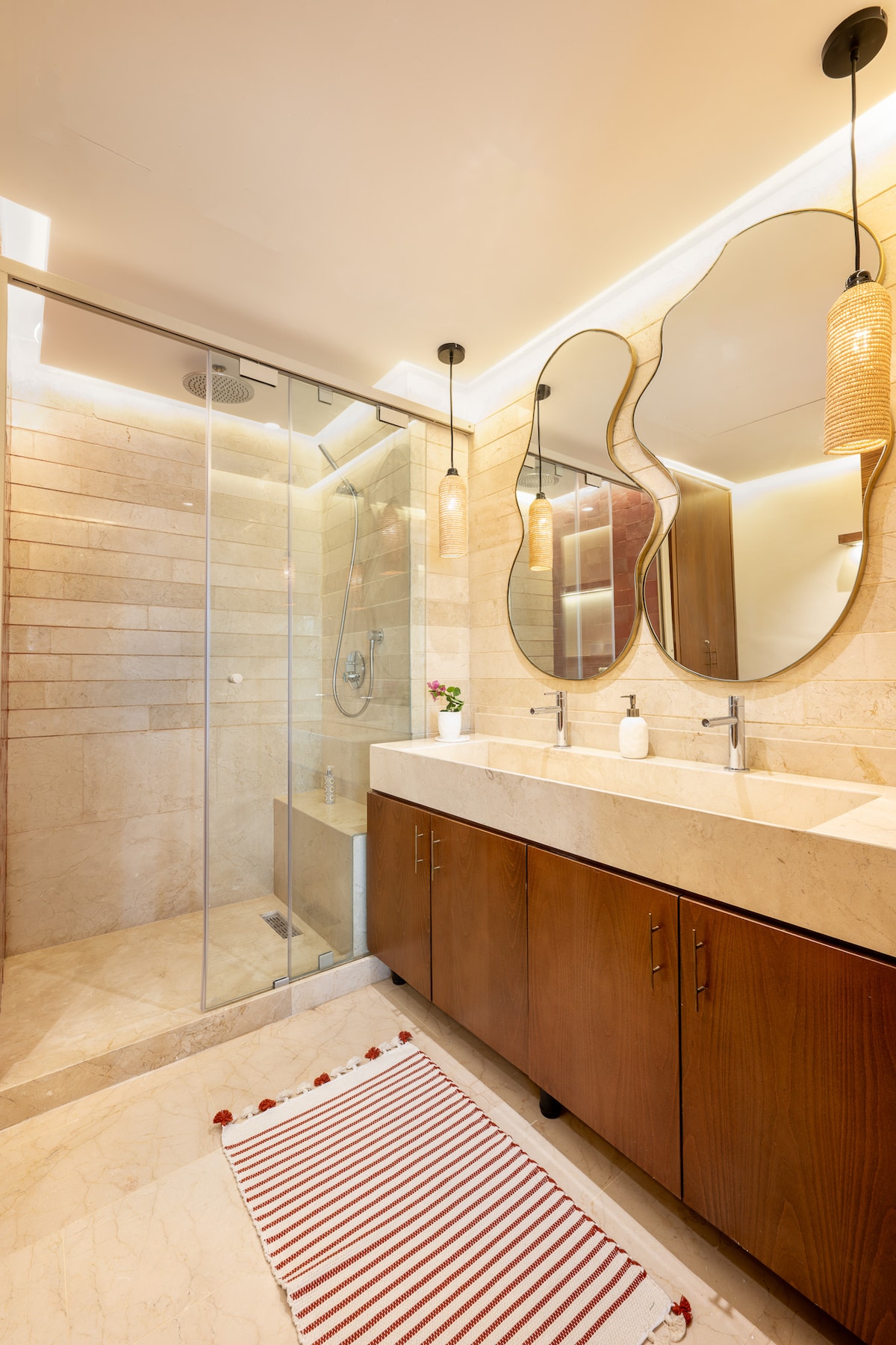 The modern bathroom features a spacious walk-in shower with a glass enclosure and a stylish double marble vanity. Decorative lighting hangs above two mirrors, enhancing the warm tones of the wooden cabinetry. A striped mat lies on the floor, adding a touch of comfort.