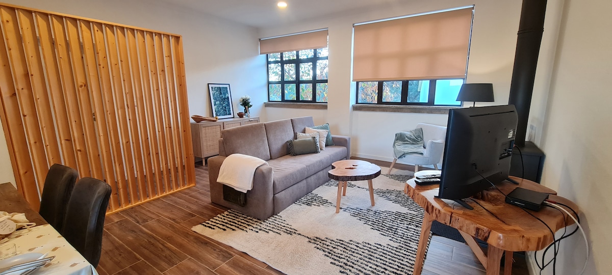 A spacious living area features a comfortable sofa, accent chairs, and a coffee table set on a patterned rug. Large windows allow natural light to brighten the room, while a wood stove adds warmth. A dining table is set for meals, enhancing the inviting ambiance.