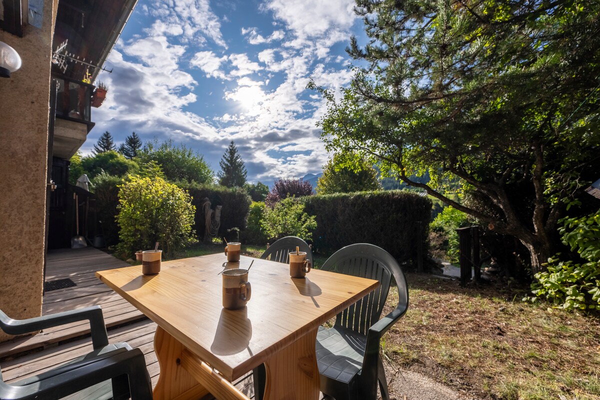 A wooden table is set on a patio, surrounded by green chairs. The space is bathed in natural light, with a cloudy sky and trees visible in the background. Lush greenery frames the garden area, creating a serene outdoor environment.