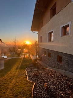 A serene sunset is visible beyond the charming farmhouse, with golden rays illuminating the landscape. The building features a combination of light stone and dark rock walls, framed by well-kept gardens and a winding pathway leading to inviting outdoor seating.