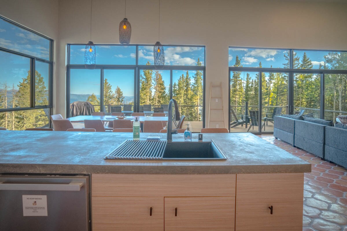 A modern kitchen is displayed with a central island featuring a sink and sleek cabinetry. Expansive windows frame a scenic view of surrounding trees and mountains, allowing natural light to fill the space. A dining area is visible in the background with additional seating.