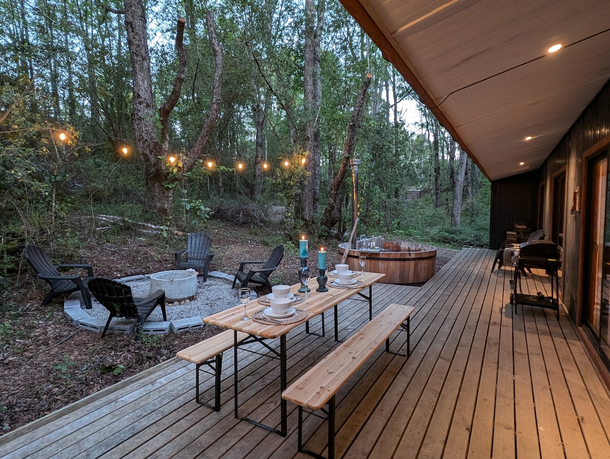 An outdoor dining area features a long wooden table set with plates and candles, surrounded by several chairs. A cozy fire pit with stone seating is seen nearby, and a wooden hot tub is positioned on the deck, complemented by string lights along the railing.