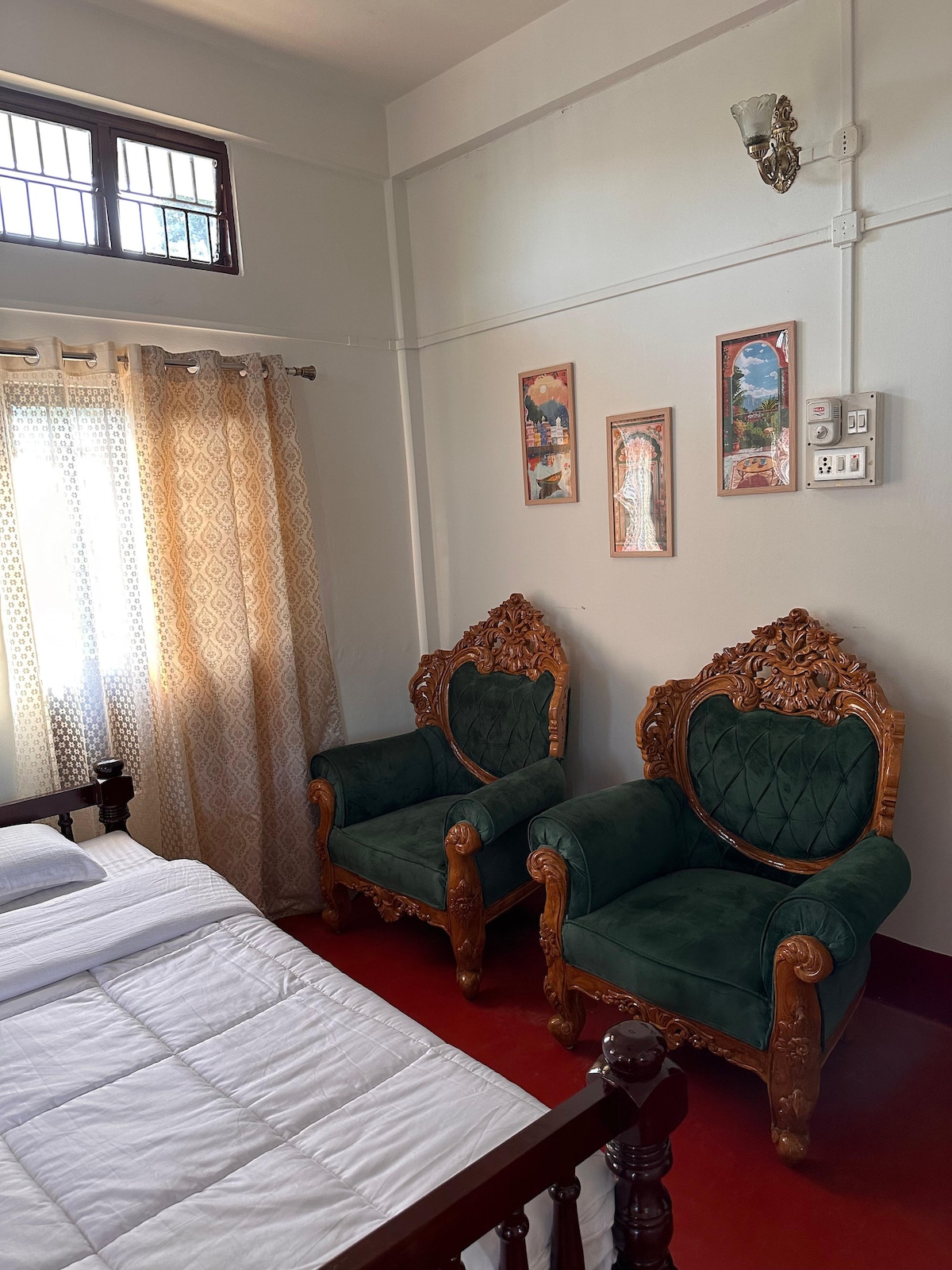 A cozy bedroom setting features a spacious bed with a white blanket and two intricately carved wooden chairs upholstered in green fabric. A pair of framed artworks hangs on the wall beside the chairs. Natural light filters through sheer curtains, creating a bright atmosphere.