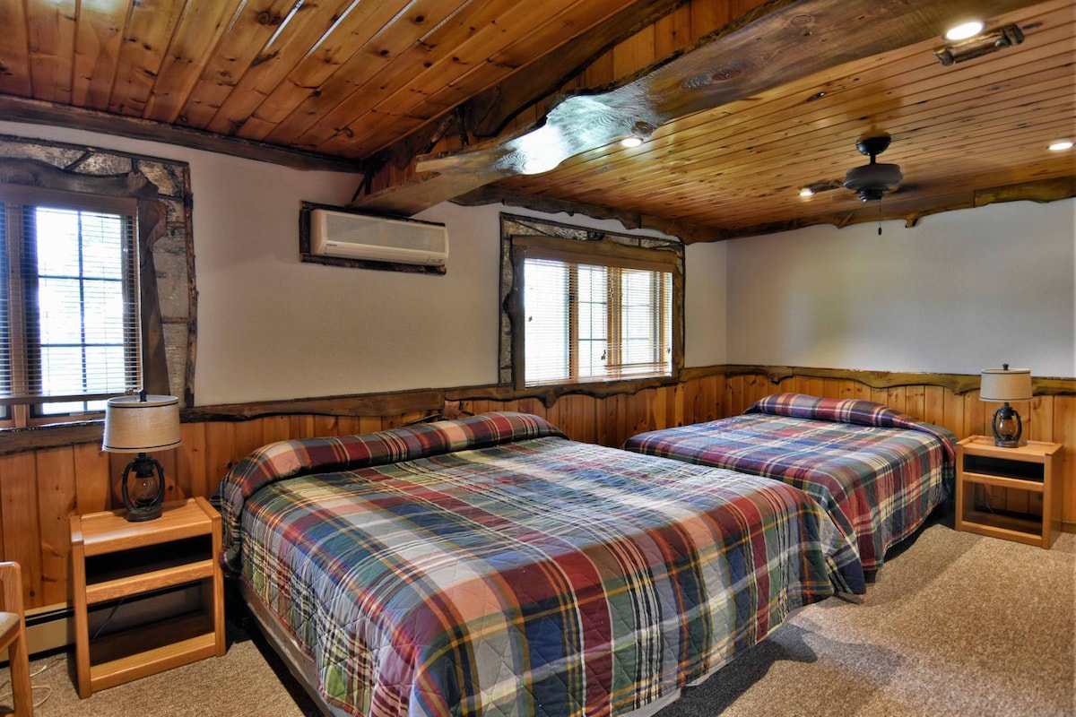 A spacious guest room features two beds dressed in plaid bedding. Wooden paneling and a beamed ceiling add a rustic touch. Natural light enters through multiple windows, complemented by bedside lamps on wooden nightstands. An air conditioning unit is mounted above the beds for comfort.