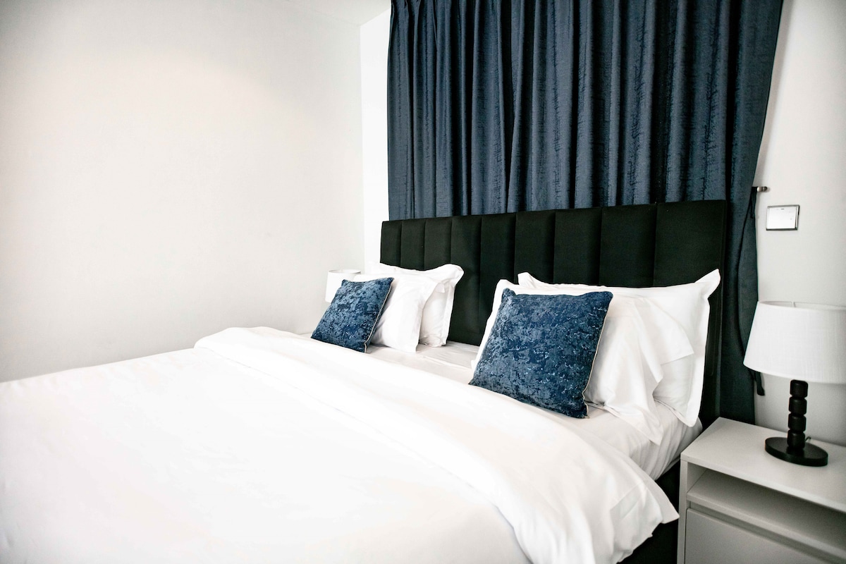 A well-appointed sleeping area features a spacious bed with crisp white linens. Two navy blue accent pillows add a touch of color, while the elegant headboard creates a sophisticated backdrop. Natural light illuminates the room, complemented by soft, dark curtains and a bedside lamp.