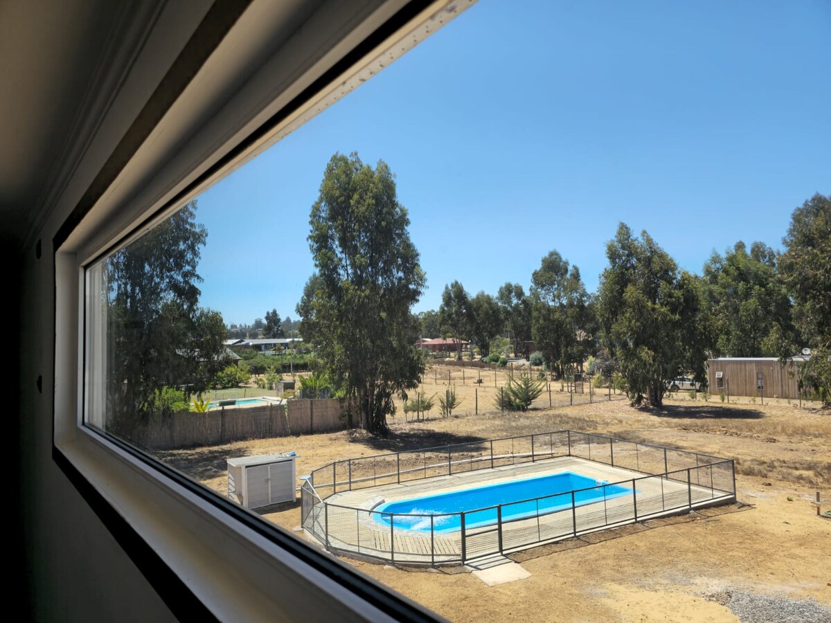 The view from the window showcases a fenced swimming pool situated in a sunlit outdoor area, surrounded by trees and open space. The landscape includes grassy patches and a distant view of other properties, emphasizing the tranquility of the surroundings.