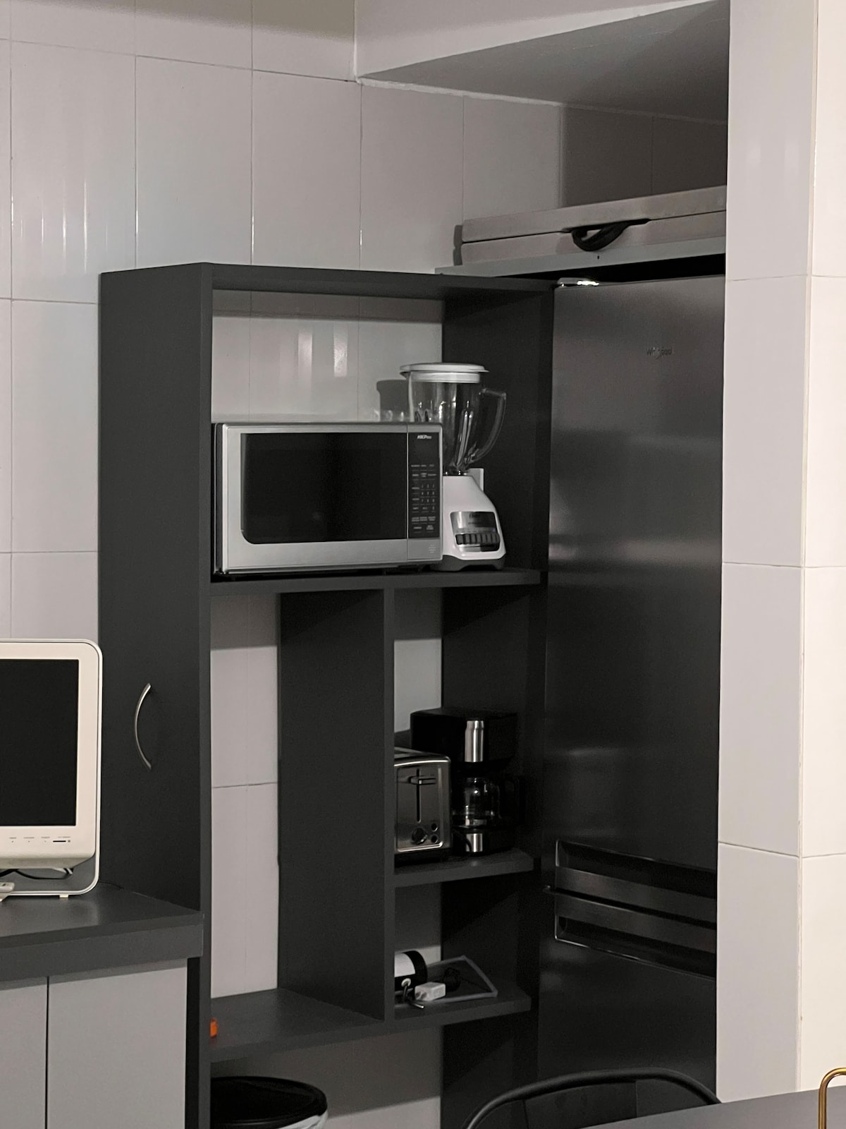A sleek kitchen setup includes a modern microwave, blender, and coffee maker placed on a dark shelving unit. A stainless steel refrigerator stands adjacent, while a computer monitor is positioned on a countertop below. The setting features tiled walls in a light color.