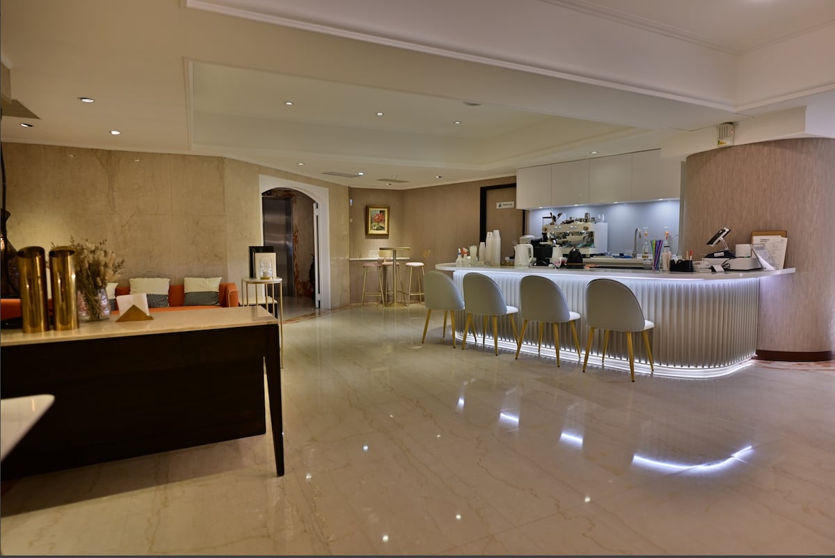 The interior of the café features a polished marble floor and softly lit walls. A sleek bar area is visible, with modern stools arranged around it. Tables are placed strategically, providing an inviting space for guests to relax and enjoy refreshments.