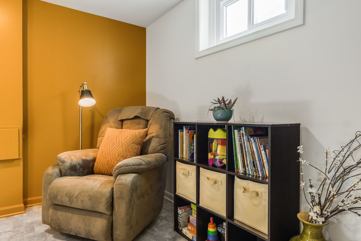 A cozy corner in the finished basement features a plush armchair, accented by a decorative orange pillow. A small bookshelf is filled with various books and games, while a light stands nearby, illuminating the space. A small potted plant adds a touch of greenery.
