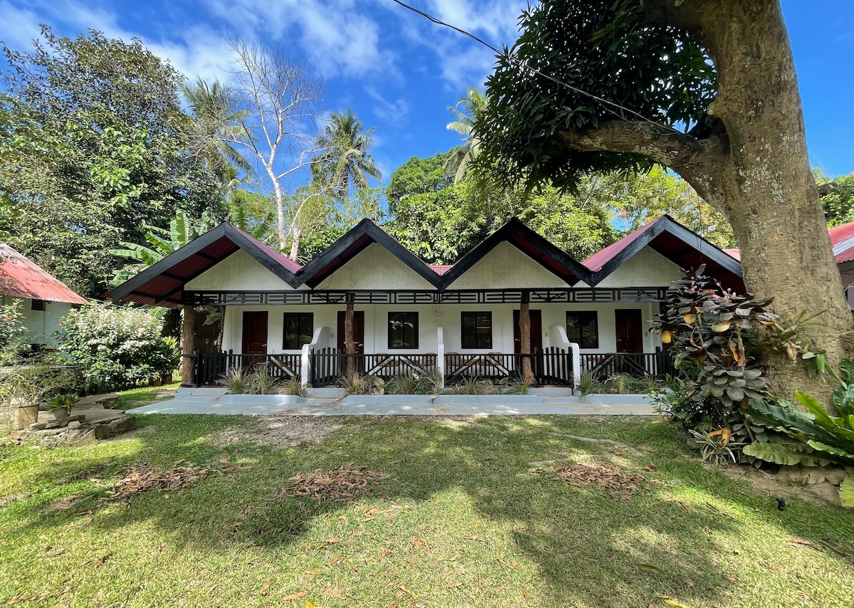A simple guesthouse is depicted, featuring a row of three private room entrances under a gabled roof. Each room is flanked by a small balcony, and a lush green lawn and tropical plants enhance the natural setting. Trees and blue skies are visible in the background.