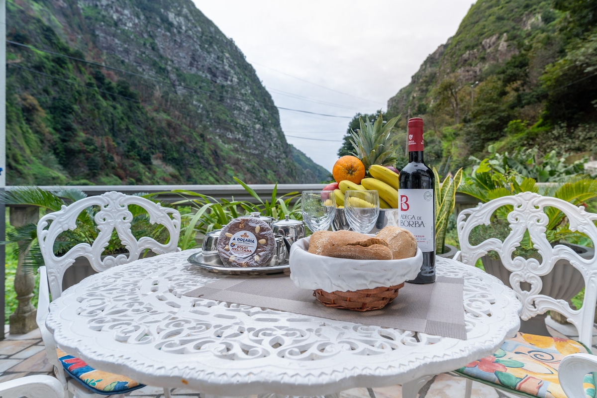 An outdoor dining table is set against a backdrop of steep green mountains. The table features a white lace design, along with a bottle of wine, glasses, a basket of bread, and a variety of fruits. Natural light enhances the serene atmosphere.